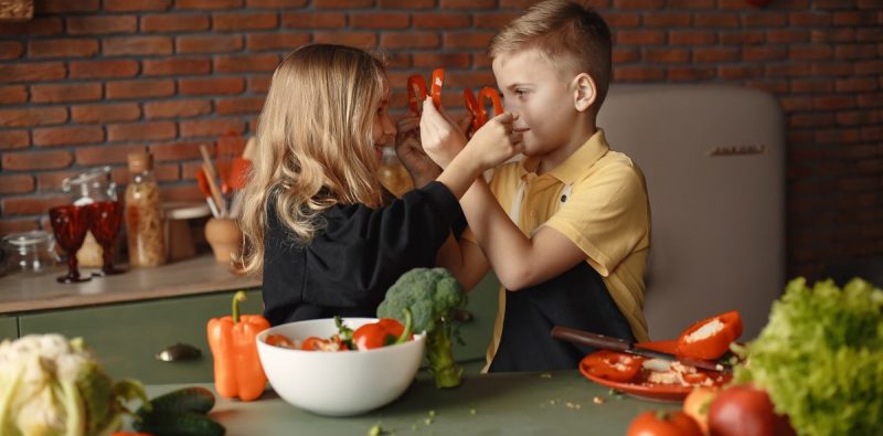 Playful boy and girl wearing apron preparing healthy vegetarian salad together at table with ingredients and bowls while looking at each other through slices of pepper